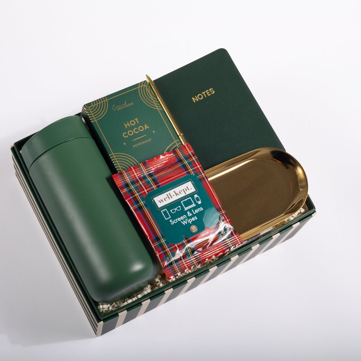 A curated gift set in a dark green striped box, featuring a dark green insulated bottle, a green notebook with gold lettering, a box of hot cocoa, a red tartan-patterned packet of Well-Apied hand sanitizer, and a gold-colored tray, all arranged on a white background.