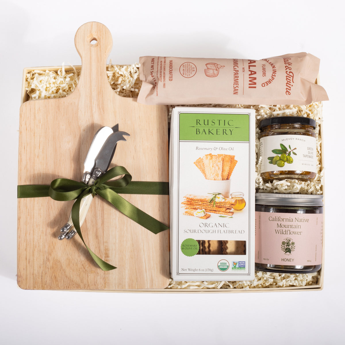 A gift hamper set on a white background, featuring a light wood cutting board with a green ribbon, a multi-tool, a box of Rustic Bakery organic sandwich bread, jars of California Native Mountain Wild Rose honey and olive oil, and a bag of Calamari.