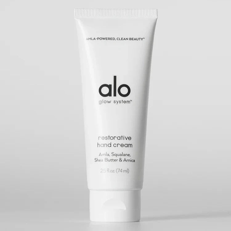 A white tube of "Alo Glow System Restorative Hand Cream" standing upright against a plain light background. The label highlights key ingredients such as amla, squalane, shea butter, and arnica, promoting it as part of a clean beauty regimen.