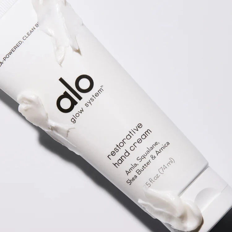 A tube of "Alo Glow System Restorative Hand Cream" laid on its side, with small amounts of cream smeared on the tube and a dollop emerging from the cap. The image highlights the product's creamy consistency and luxurious feel.