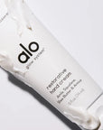 A tube of "Alo Glow System Restorative Hand Cream" laid on its side, with small amounts of cream smeared on the tube and a dollop emerging from the cap. The image highlights the product's creamy consistency and luxurious feel.