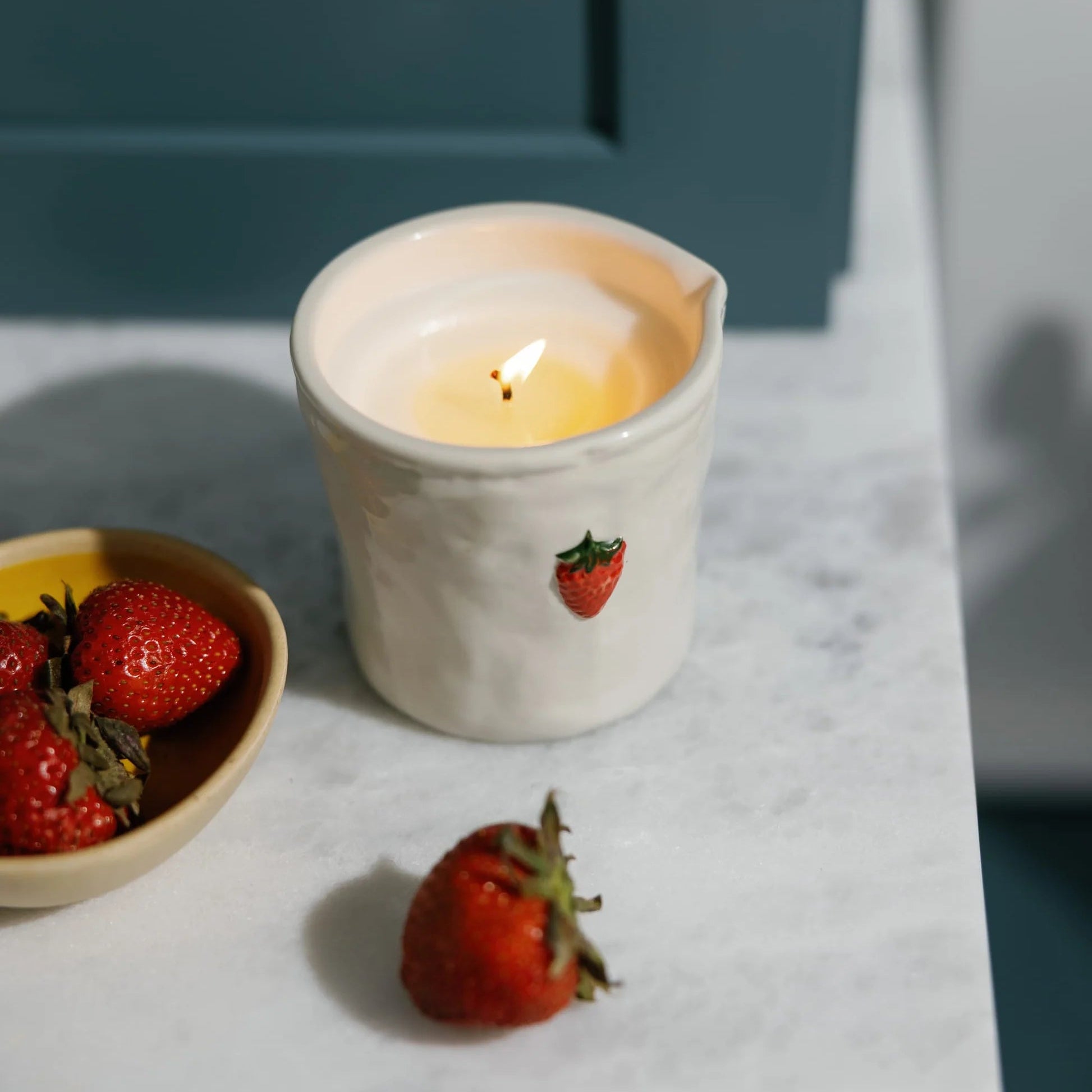 A lit cream-colored ceramic candle holder with a small red strawberry illustration, placed on a white marble surface next to a bowl of fresh strawberries.