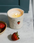 A lit cream-colored ceramic candle holder with a small red strawberry illustration, placed on a white marble surface next to a bowl of fresh strawberries.