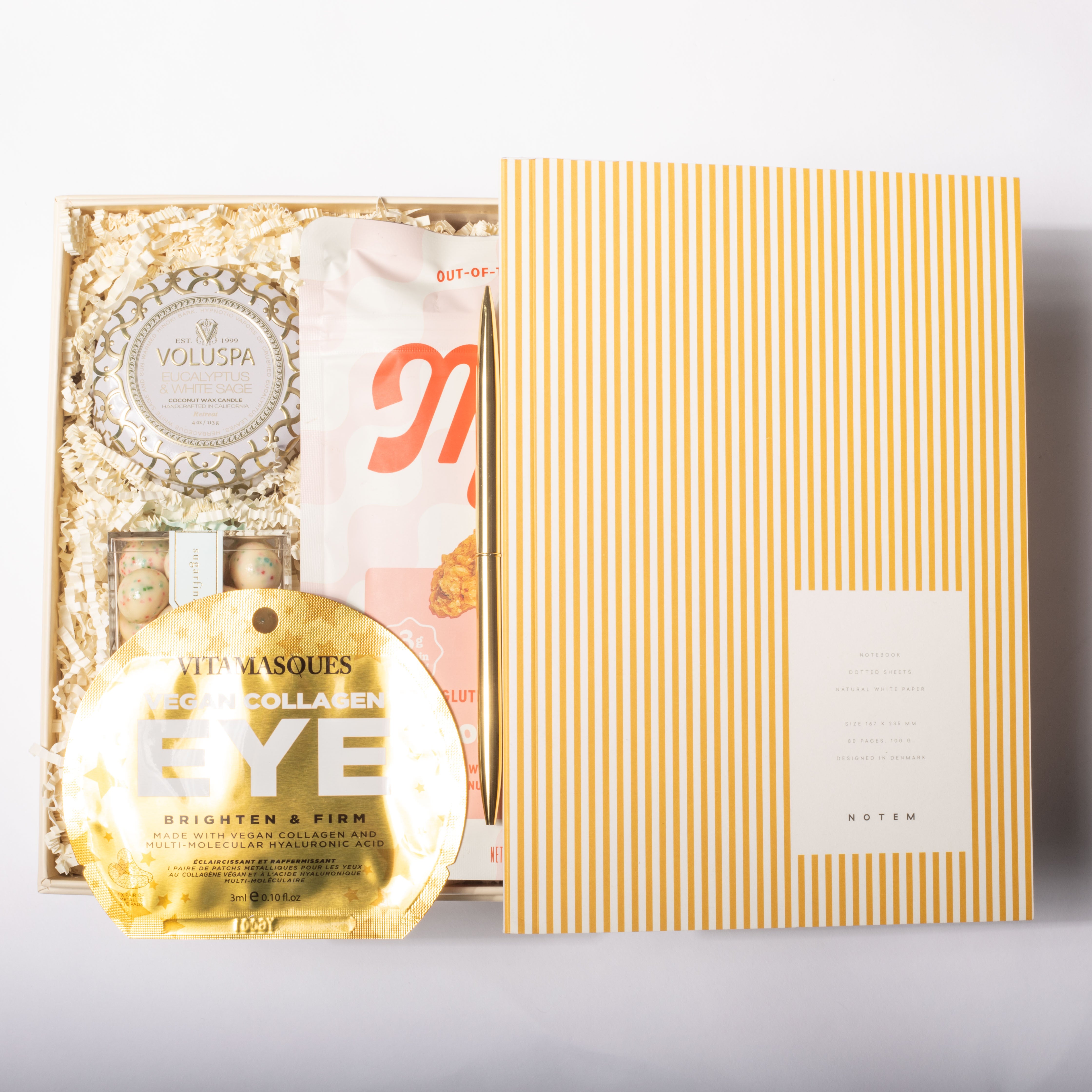 A curated beauty gift set arranged on a white background, featuring a gold Notem envelope, a gold round eye mask, a white and pink pouch, and a boxed set with shredded paper filler and a round Voluspa item.