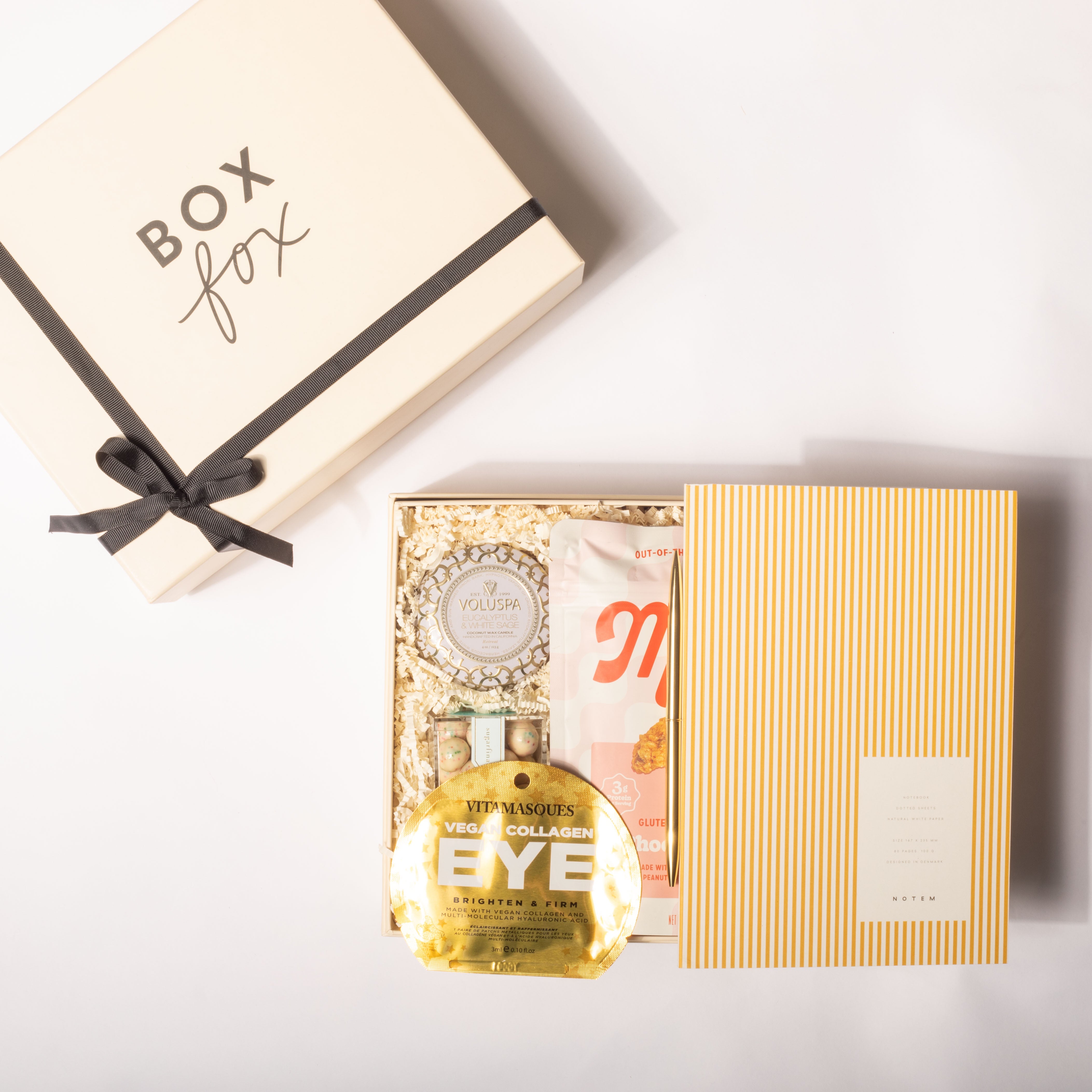 A curated gift box set from Box Box, featuring a cream-colored outer box with a black ribbon, and an open inner box containing a gold bottle of eye cream, a pink and white pouch, and a yellow-and-white striped box, all arranged on a white background.