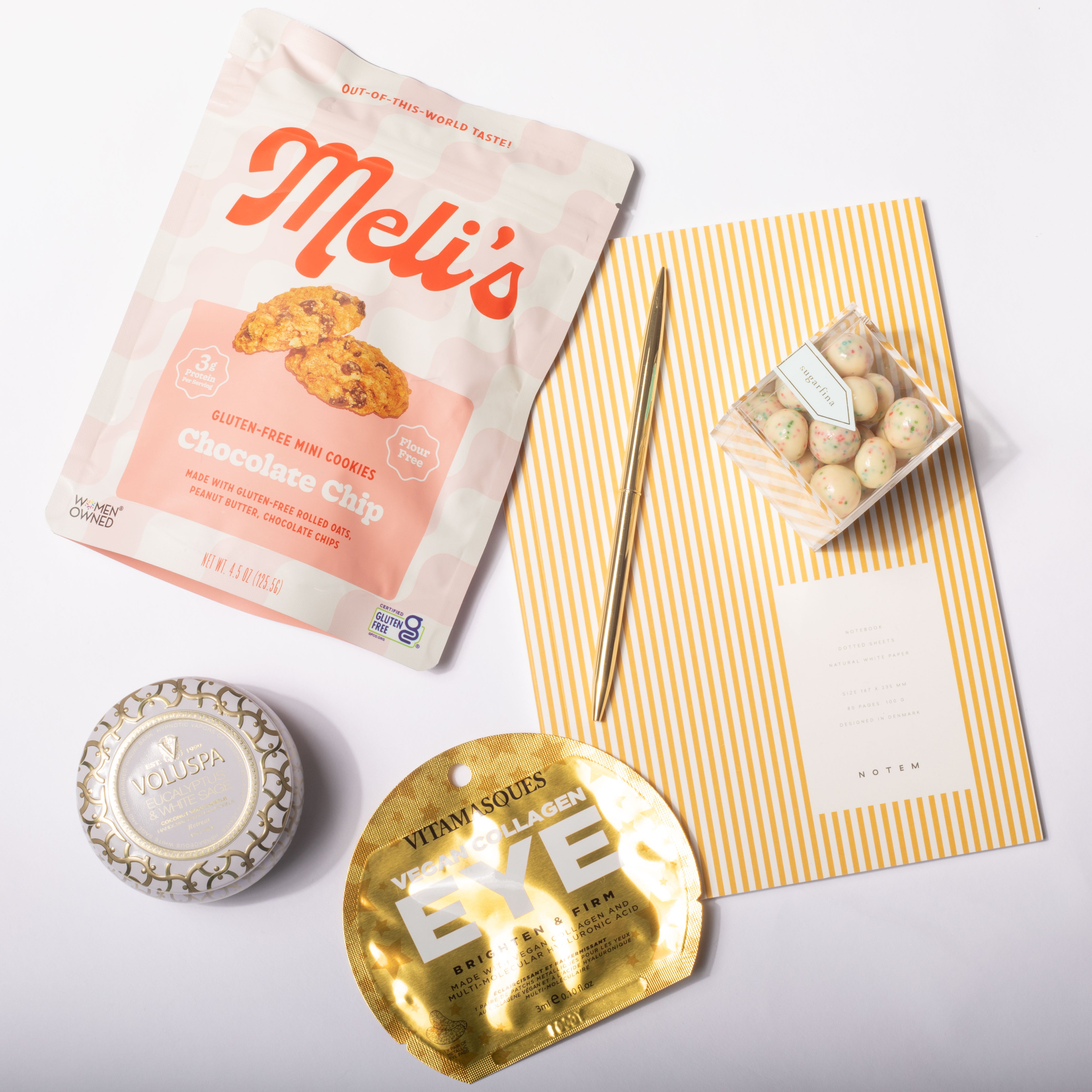 A curated flat-lay of beauty and snack items on a white background, including a bag of Meli's chocolate chip cookies, a small tin of gold eyeshadow, a gold eyelash pack, and a yellow-and-white striped card with a small wrapped treat.