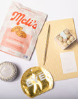 A curated flat-lay of beauty and snack items on a white background, including a bag of Meli's chocolate chip cookies, a small tin of gold eyeshadow, a gold eyelash pack, and a yellow-and-white striped card with a small wrapped treat.