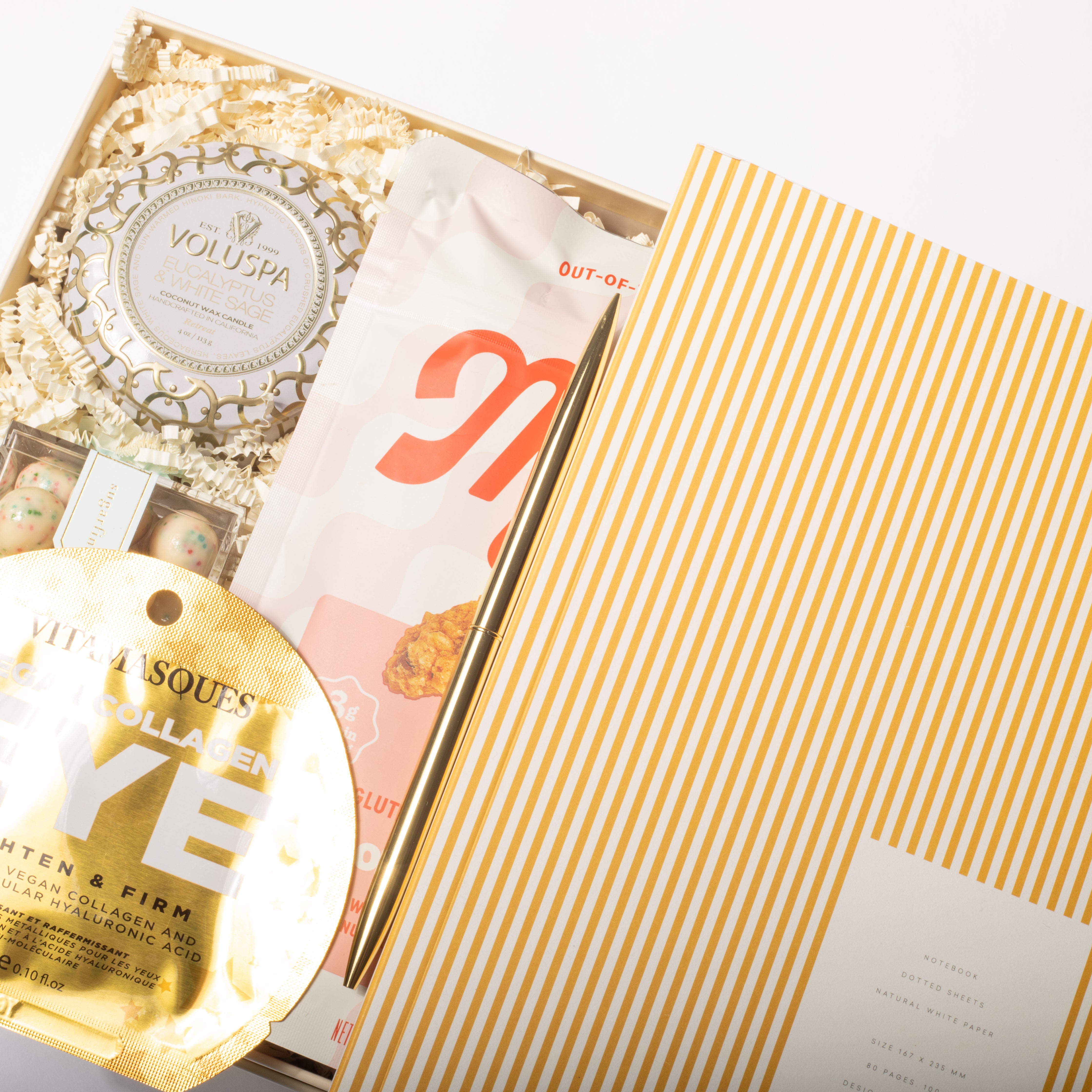 An open gift box containing a round gold collagen face mask packet, a white and gold Voluspa wax mask tin, and a pink and white cosmetic pouch, with a yellow-and-white striped outer box partially visible. The products are arranged on a white background.