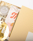 An open gift box containing a round gold collagen face mask packet, a white and gold Voluspa wax mask tin, and a pink and white cosmetic pouch, with a yellow-and-white striped outer box partially visible. The products are arranged on a white background.