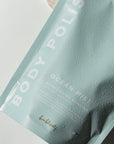 Rectangular, sealed, light blue plastic bag with white text that reads "BODY POLISH" on the side and "OCEAN MIST MOISTURIZING EXFOLIANT" in bottom right. Photographed on grey background.