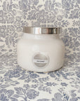 White glass jar candle with silver lid on a blue and white floral backdrop.