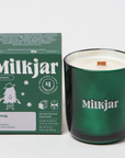 The image shows a green glass candle with a matching green box beside it. The box and candle both display the brand name "Milkjar." The box is labeled with the scent name "Doug," and lists fragrance notes: evergreen, bourbon, and musk. The label mentions that the candle is made of coconut soy wax and that $1 from each candle is donated to charity. The candle has a wooden wick visible at the center.