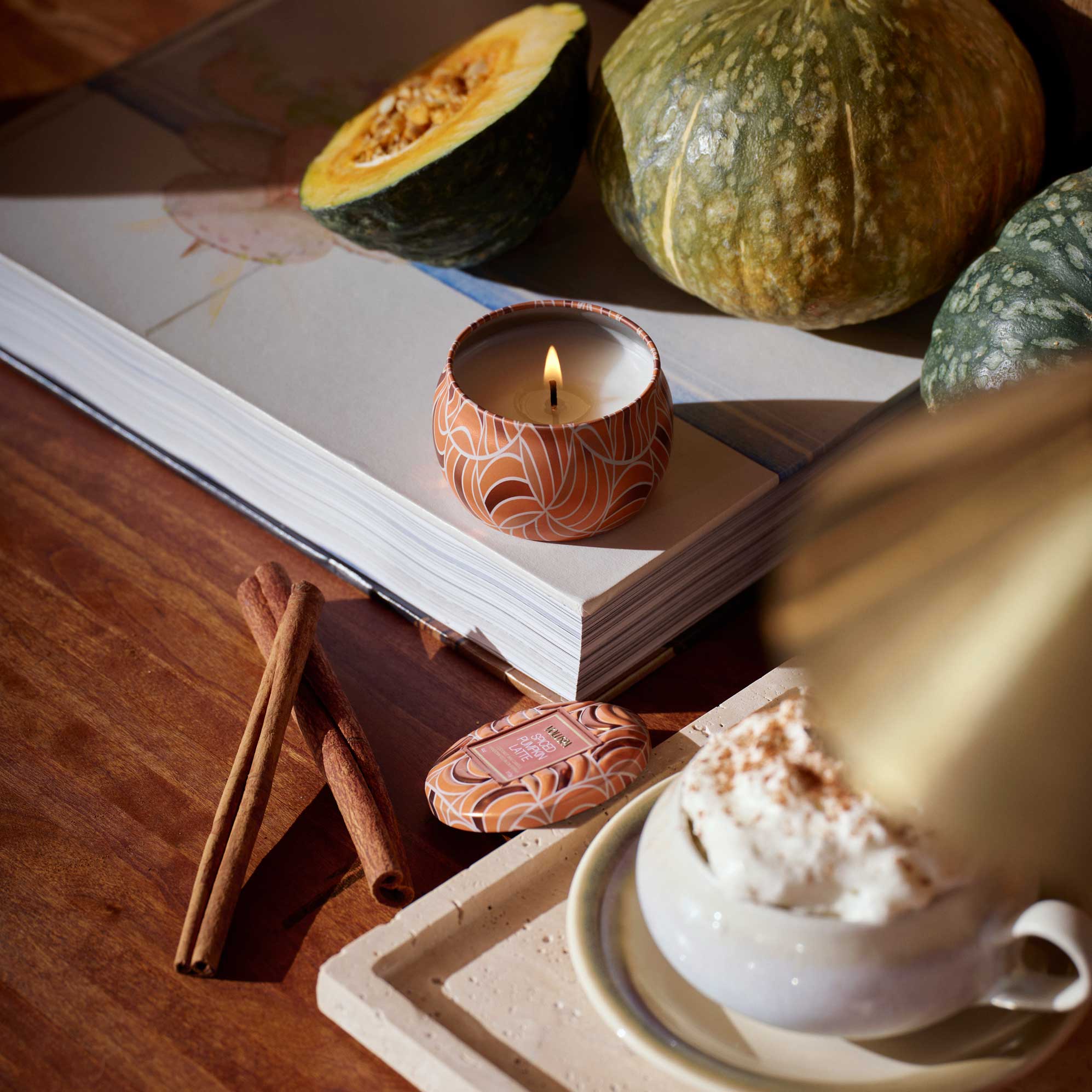 Candle with decorative container on an open book, surrounded by pumpkins, a cinnamon stick, and a cup of coffee on a wooden surface.
