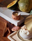 Candle with decorative container on an open book, surrounded by pumpkins, a cinnamon stick, and a cup of coffee on a wooden surface.