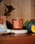 Candle with decorative packaging on a table with pumpkins and a dessert.
