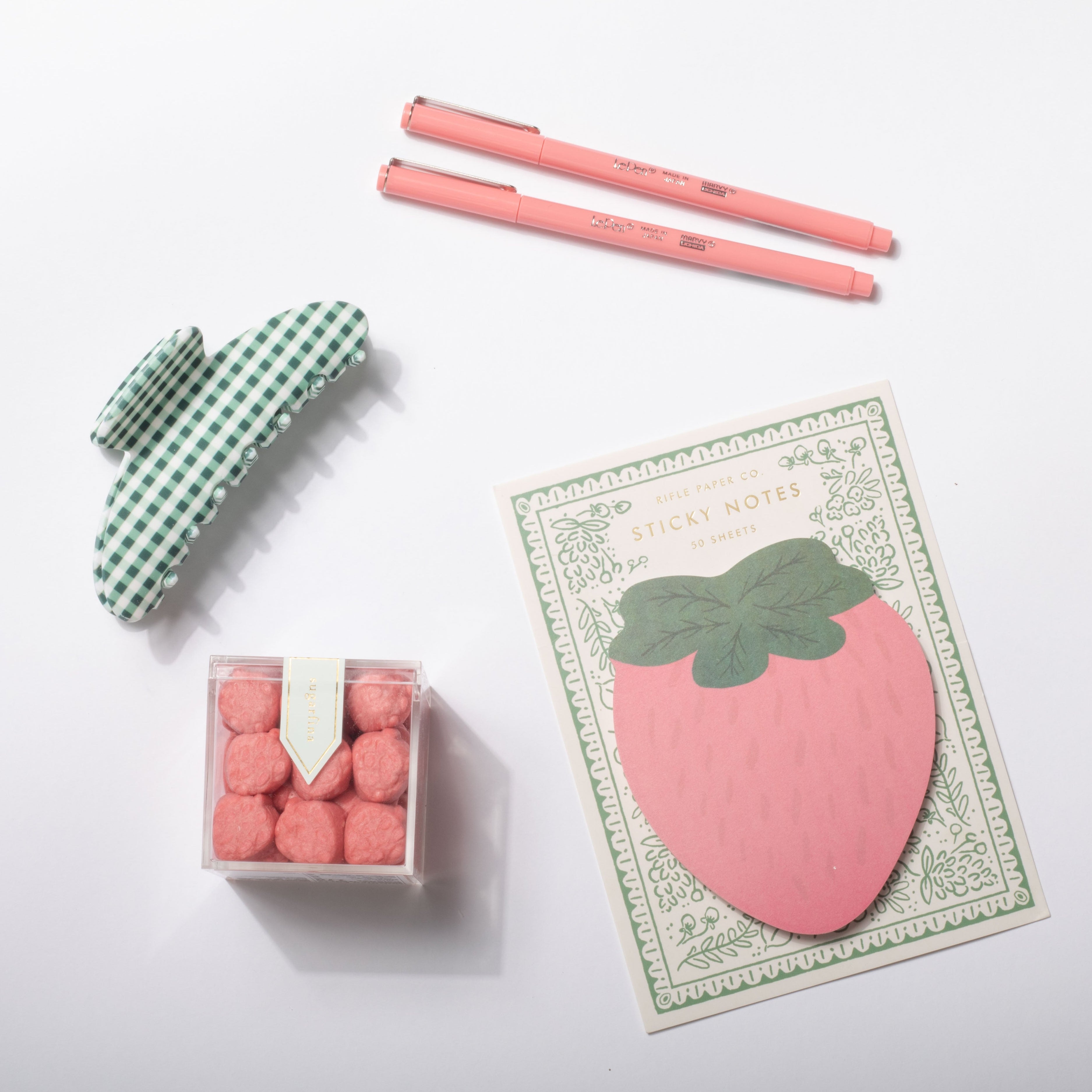 Set of stationery items including a pink strawberry-shaped post-it note, two pink pens, a green and white checkered eraser, and a small box of pink pom-poms on a white background.
