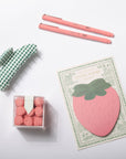 Set of stationery items including a pink strawberry-shaped post-it note, two pink pens, a green and white checkered eraser, and a small box of pink pom-poms on a white background.