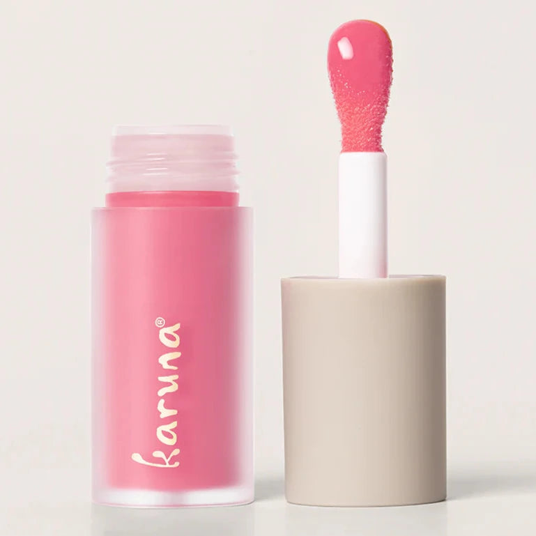 A pink colored lip oil that reads, "KARUNA." The doe-foot applicator is displayed next to the lip oil.