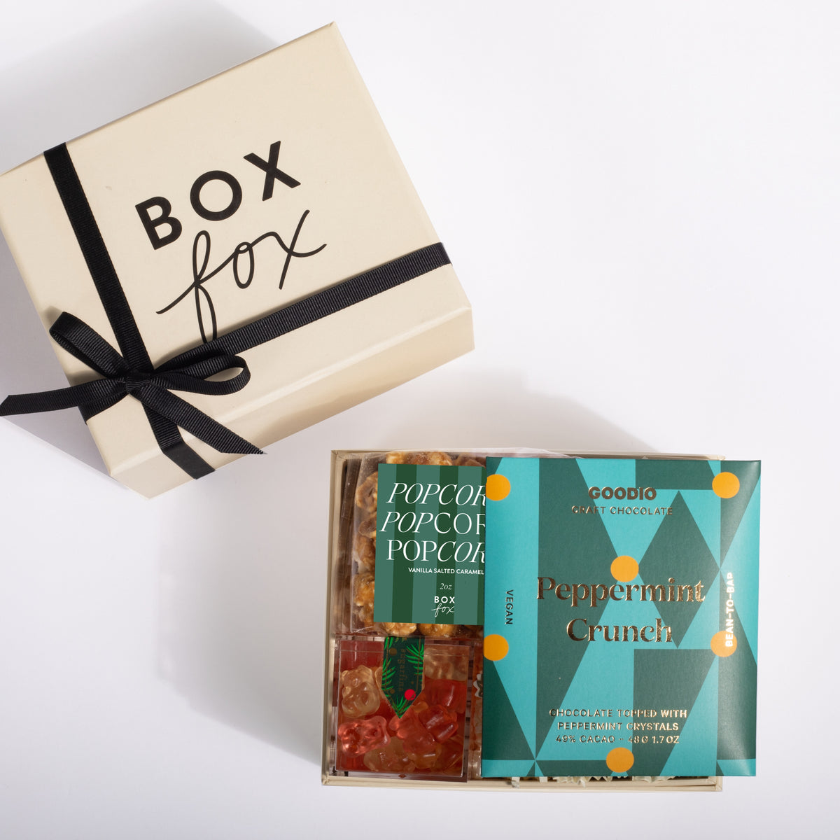 A gourmet popcorn gift box set from Goodoio by Paccola, featuring a cream-colored gift box with a black ribbon and a teal 'Peppermint Crunch' box containing two varieties of popcorn, displayed on a clean white background.
