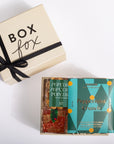 A gourmet popcorn gift box set from Goodoio by Paccola, featuring a cream-colored gift box with a black ribbon and a teal 'Peppermint Crunch' box containing two varieties of popcorn, displayed on a clean white background.