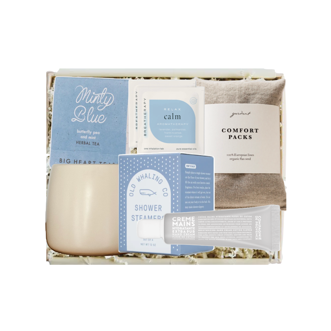 A curated self-care gift box set from Big Hearted Things, featuring a minty blue herbal tea packet, a calming bath soak packet, a shower steam packet, a comfort pack, a tube of natural nail care, and a beige ceramic candle, all arranged in a white gift box with shredded paper filler.
