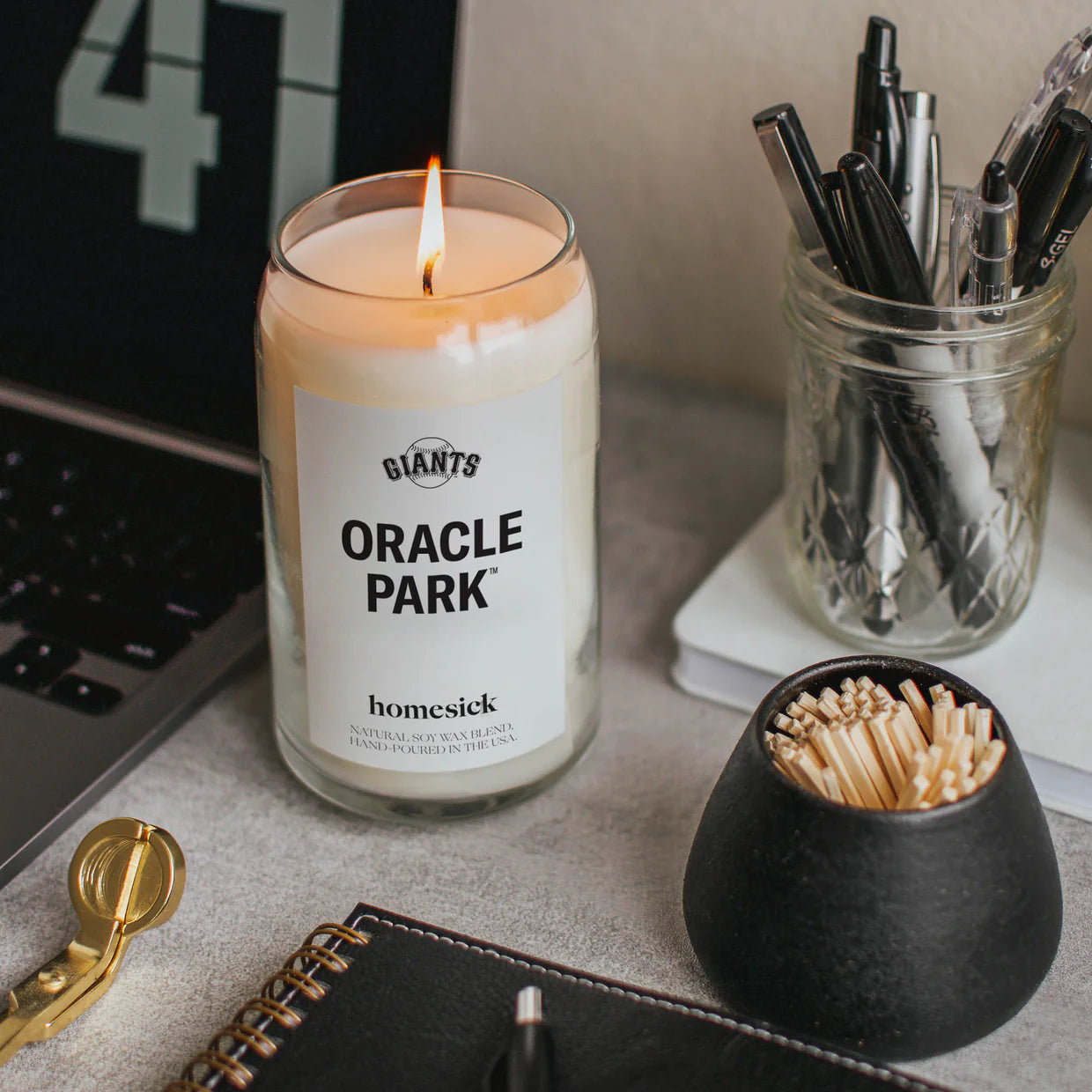 Oracle Park Candle on desk next to computer, matches, pens and notebook