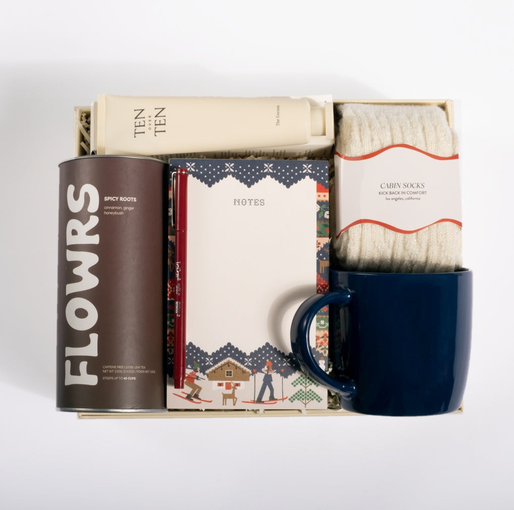 A curated gift box set containing a dark brown canister of 'FLOWRS' spice roots, a cream tube of 'TEN TEN' lotion, a patterned notebook, a pair of cream 'CABIN SOCKS', and a dark blue ceramic mug, all arranged in a white box on a white background.