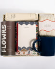 A curated gift box set containing a dark brown canister of 'FLOWRS' spice roots, a cream tube of 'TEN TEN' lotion, a patterned notebook, a pair of cream 'CABIN SOCKS', and a dark blue ceramic mug, all arranged in a white box on a white background.