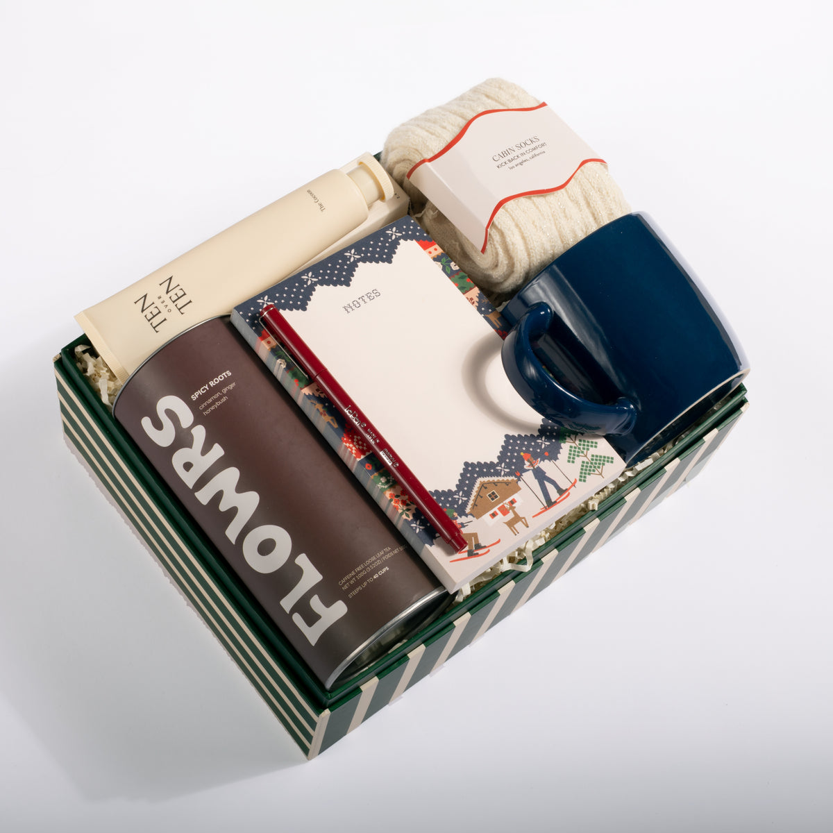 A curated gift box set containing a navy blue ceramic mug, a brown can of Flowers coffee, a cream tube of lotion, a patterned notebook, and a folded cream knit item, all arranged in a striped green and white box on a white background.