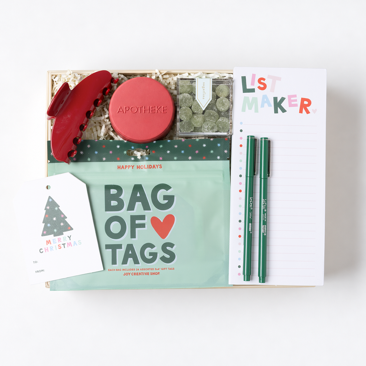 A curated gift box set on a white background, featuring a mint green 'Bag of Tags' pouch, a red hair claw clip, a round red 'Apothke' tin, a clear container of green tea bags, a white 'List Maker' notepad, and two green pens.