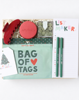 A curated gift box set on a white background, featuring a mint green 'Bag of Tags' pouch, a red hair claw clip, a round red 'Apothke' tin, a clear container of green tea bags, a white 'List Maker' notepad, and two green pens.