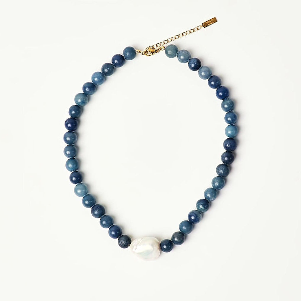 A beaded necklace featuring dark blue and lighter blue round beads, a single white pearl-like bead at the center, and a gold-tone toggle clasp, displayed on a clean white background.