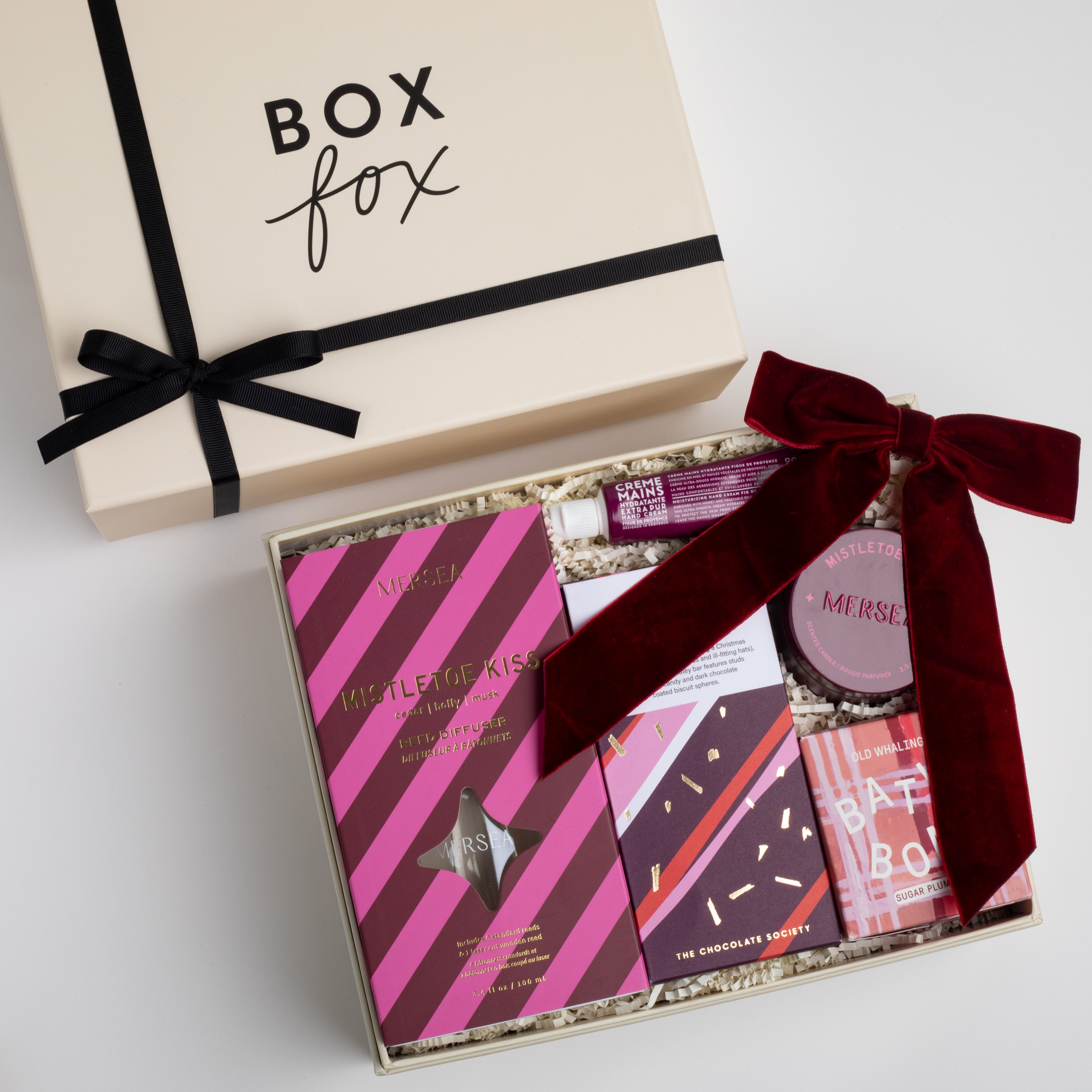 CREME BOXFOX BOX DISPLAYED OPEN WITH: Mistletoe Kiss Reed Diffuser, Mistletoe Kiss Tin Candle, Christmas Cracker Bar, Sugarplum Ferry Bath Bomb, Maroon Velvet Bow Hair Clip, Fig of Provence Travel Hand Cream