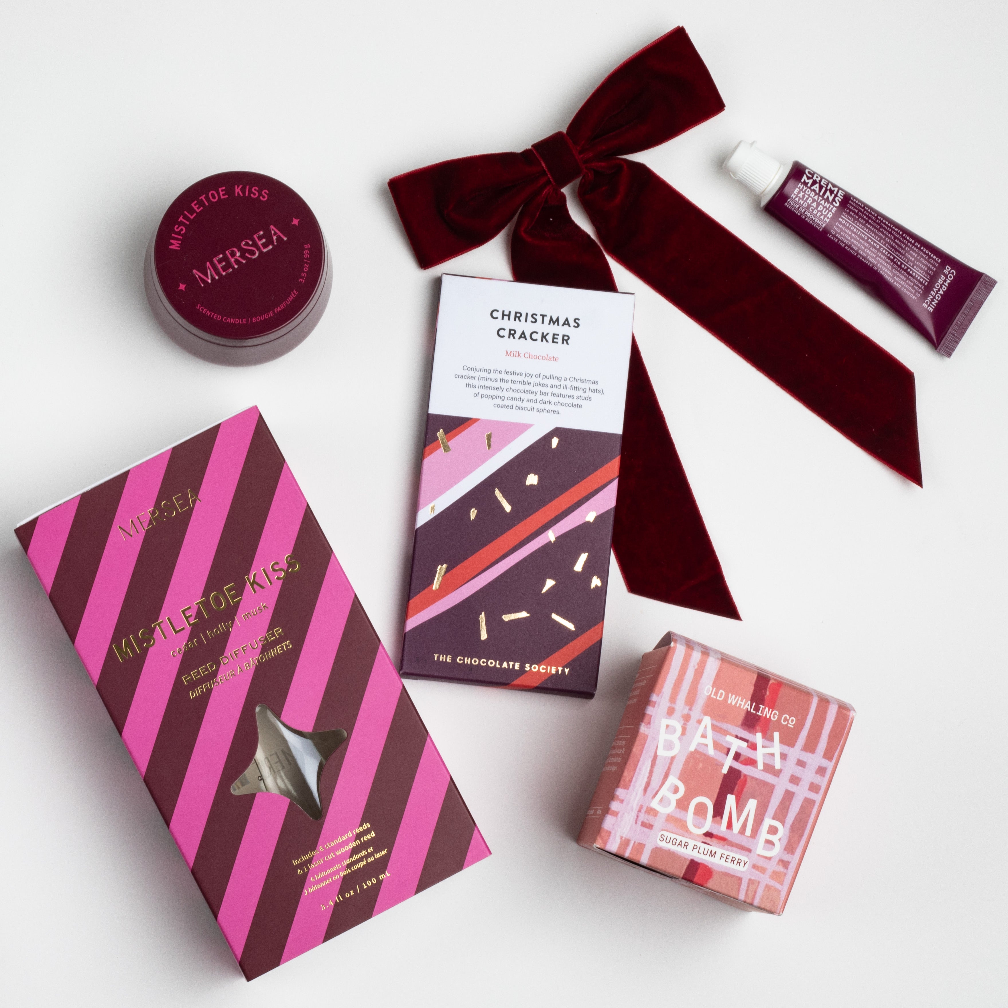 JOYEUX NOELLE CURATED BOX ITEMS DISPLAYED: Mistletoe Kiss Reed Diffuser, Mistletoe Kiss Tin Candle, Christmas Cracker Bar, Sugarplum Ferry Bath Bomb, Maroon Velvet Bow Hair Clip, Fig of Provence Travel Hand Cream