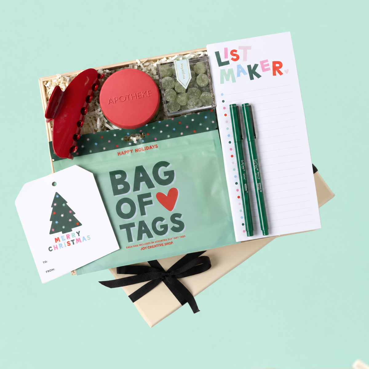A curated gift set arranged on a light mint background, including a kraft gift box with shredded paper, a red hair claw clip, a round red soap bar labeled 'APOTHEKE', a mint green 'BAG OF TAGS' pouch, a white 'LIST MAKER' notepad, two green pens, and a white Christmas greeting card tied with a black ribbon.