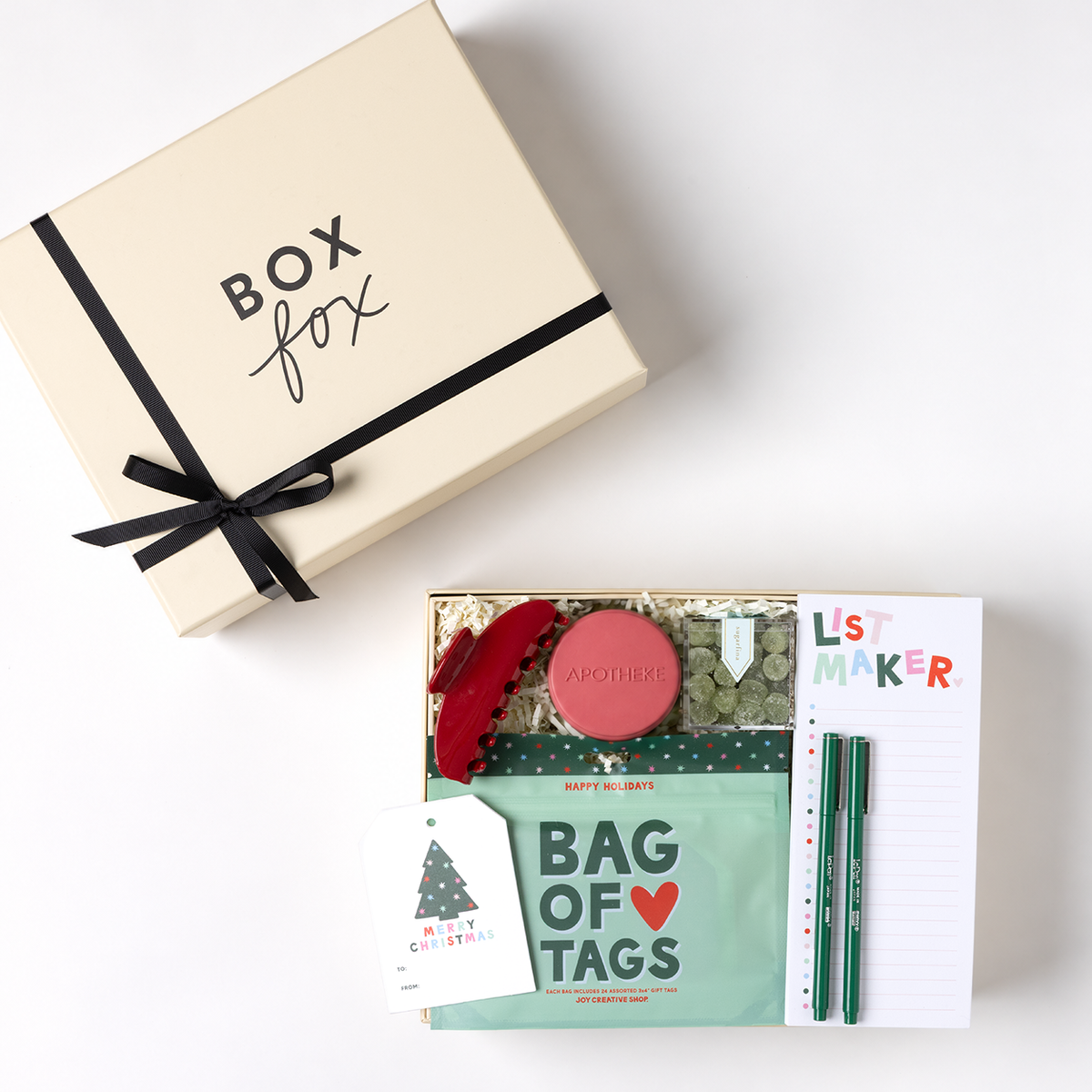 A Christmas-themed gift box set from 'Box fox' shown in a flat-lay view. The set includes a cream-colored gift box with a black ribbon, and an open box containing a mint green 'Bag of Tags' booklet, a red elastic hair tie, a pink circular item, a white 'List Maker' notepad, and two green pencils.