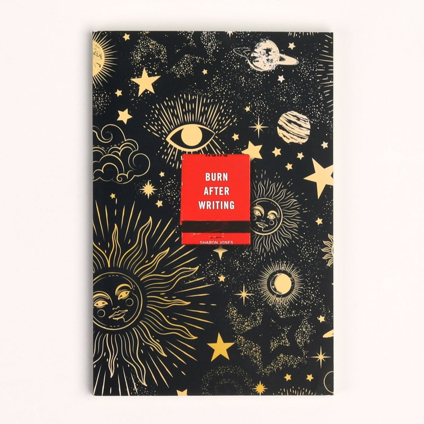Black rectangular notebook with gold embossed celestial design all over. Red matchbook in middle of book has white text that reads, &quot;Burn After Writing&quot;. Photographed on white background.