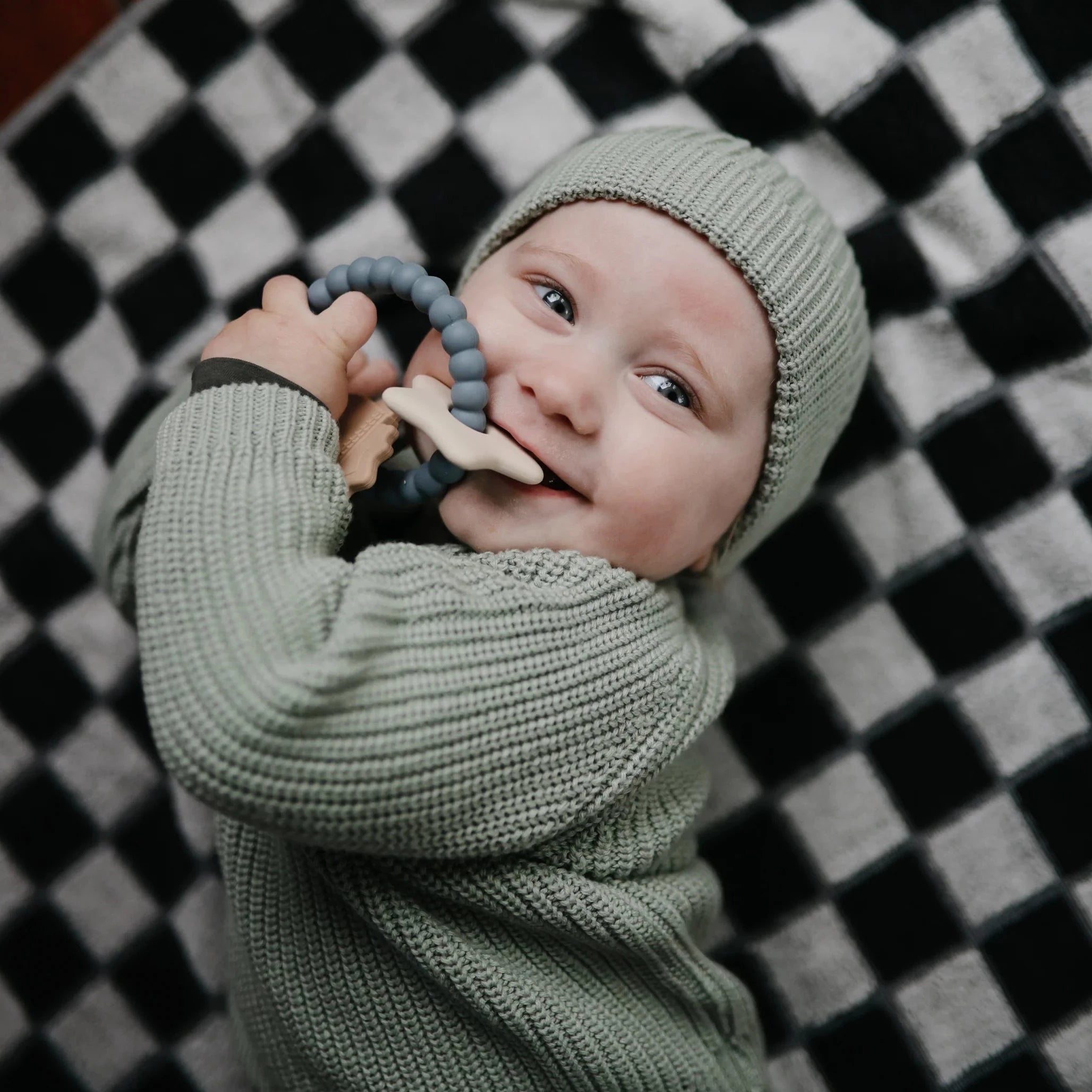 Baby holding teether with star charm in mouth. baby is wearing green hat and green knitted sweater. Baby is laying on black and white checkered blanket