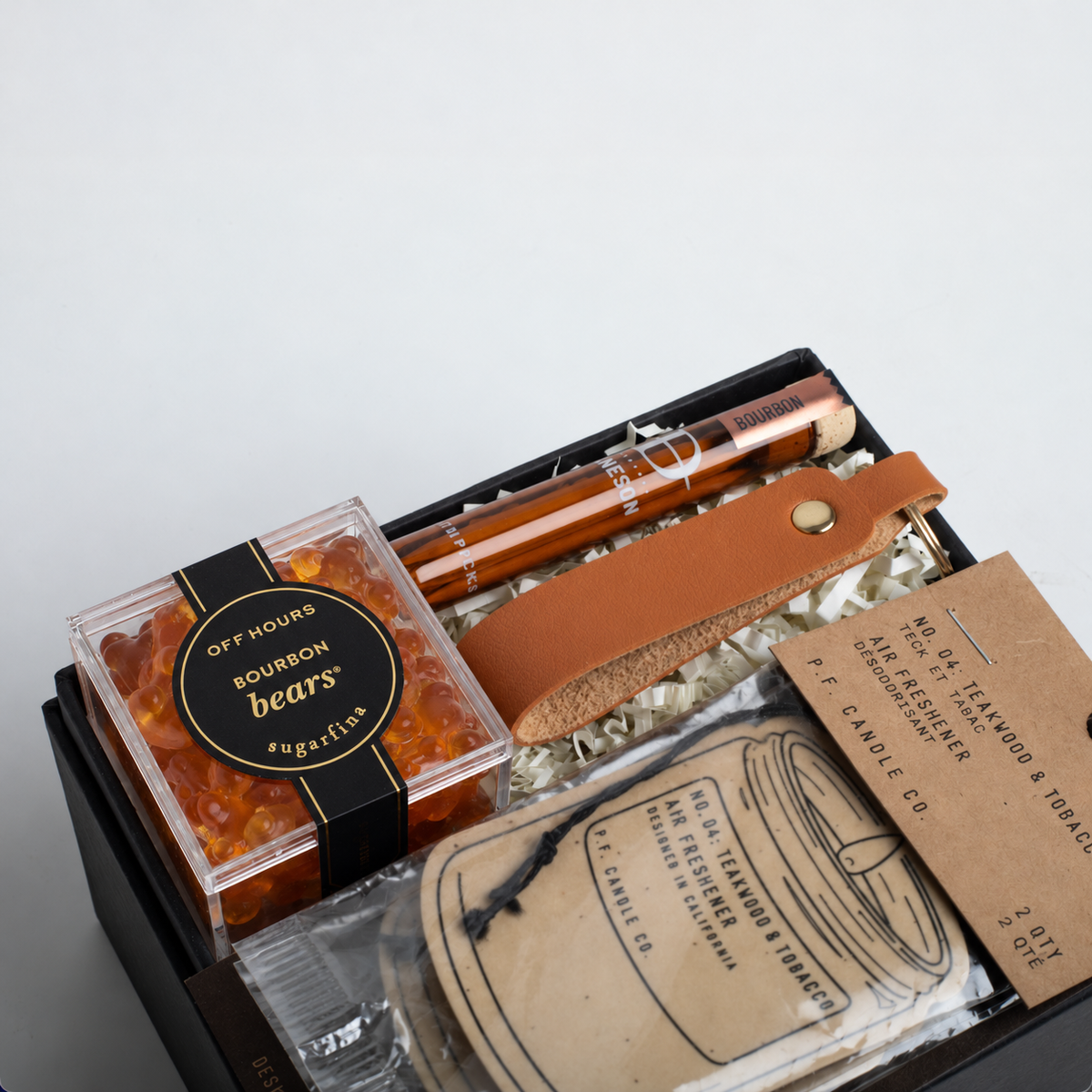A curated gift box containing a clear container of amber-colored 'Bourbon bears' wax melts, a brown leather strap, a wooden pipe, and various branded paper tags and accessories inside a black box.