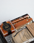A curated gift box containing a clear container of amber-colored 'Bourbon bears' wax melts, a brown leather strap, a wooden pipe, and various branded paper tags and accessories inside a black box.