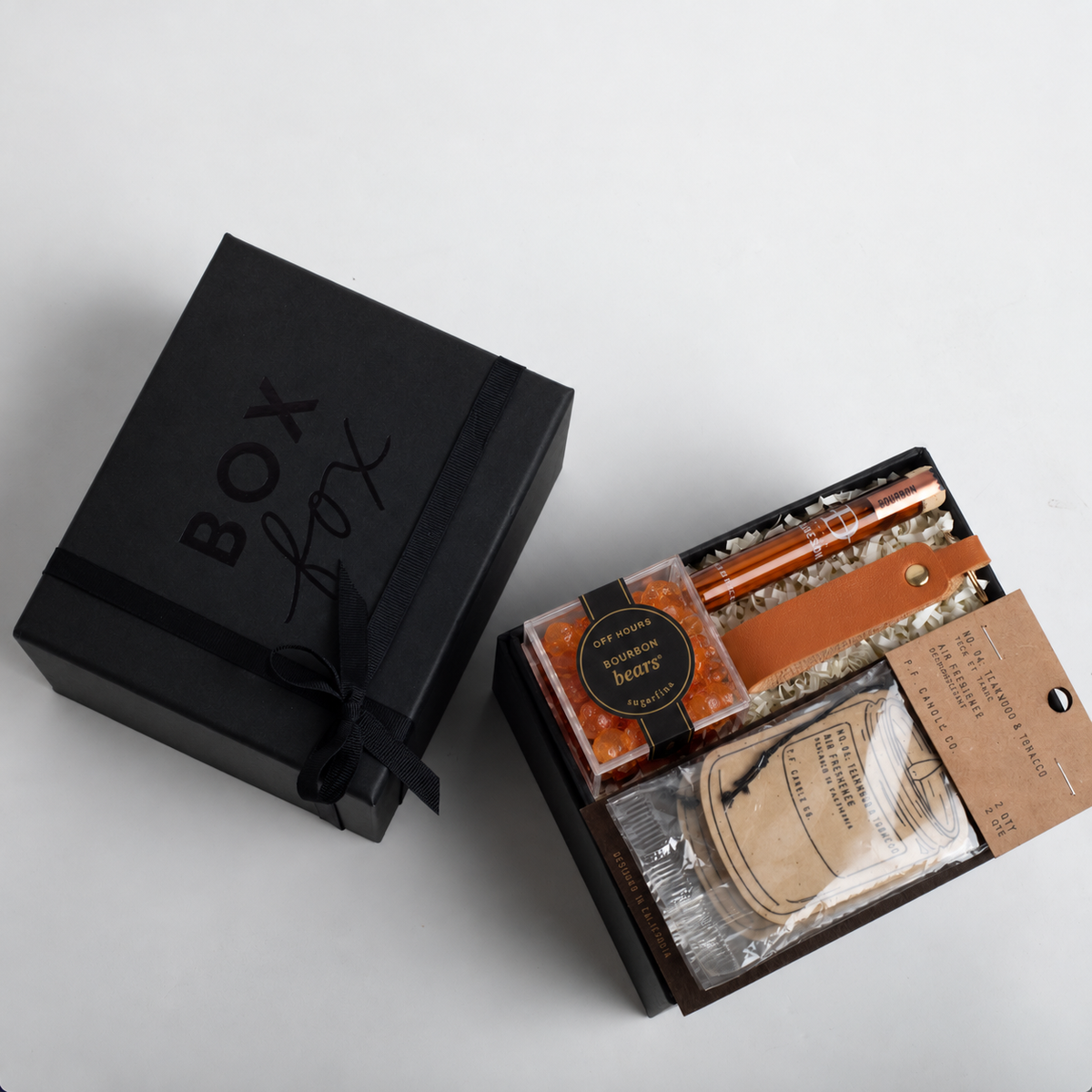 A black gift box with 'BOX box' printed on the lid sits beside an open box revealing a curated gift set with bath salts, a candle, and a packaged item on a light gray background.