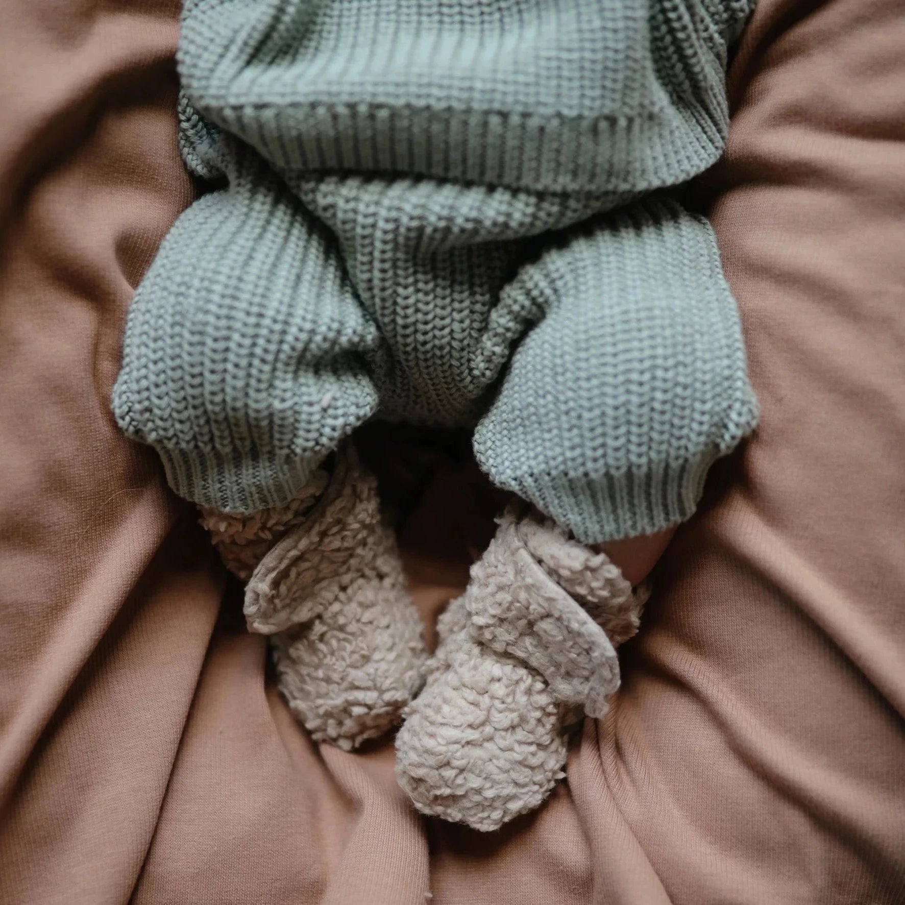 baby laying on mauve blanket with brown booties on. baby is wearing green knitted two piece set