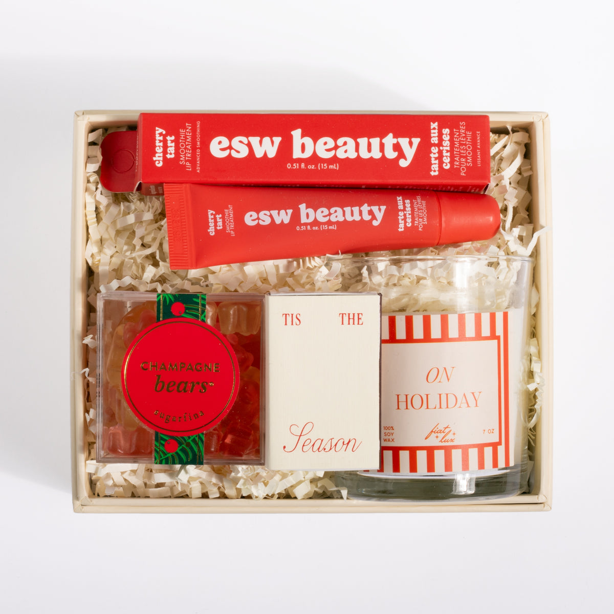 A curated gift set in a light wood box filled with shredded paper, featuring two red esw beauty lip balm tubes, a round red and green 'Champagne Bears' candle tin, a small cream-colored box with 'Tis The Season' script, and a striped red and white candle in a glass jar.