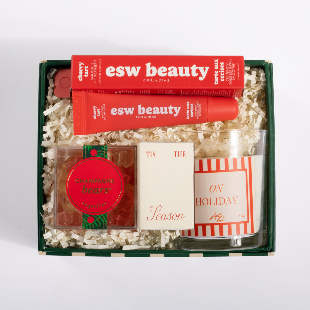 A curated gift box set containing an esw beauty lip balm tube, a red and green cherry lip balm, a candle, and a small card, all arranged in a dark green box filled with white shredded paper.