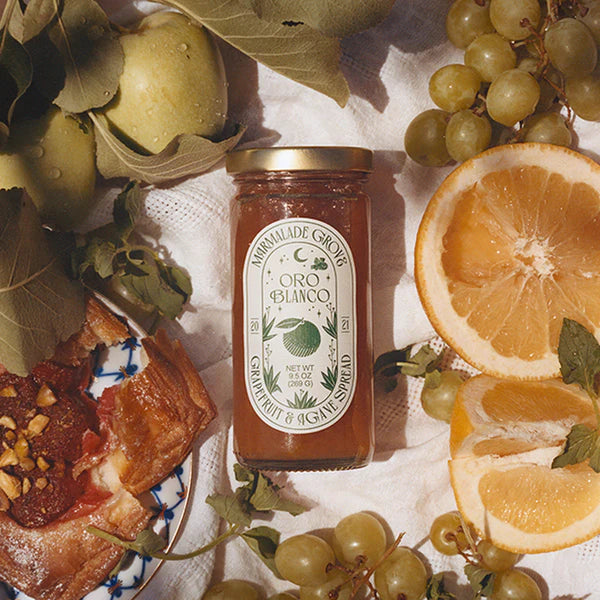 Jar of Oro Blanco grape jelly surrounded by fruits and pastries on a white cloth.