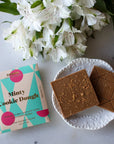 Square light brown chocolate on a ceramic white bowl. To the top of it are white flowers. Next to the bowl, to the left, is the pastel geometric packaging for the chocolate 