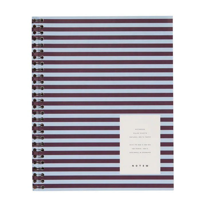 A spiral-bound notebook with horizontal dark purple and white stripes, featuring a white label on the front with the brand name 'STAN' and product details, set against a plain white background.