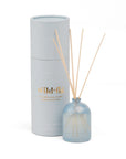 A reed diffuser set with a light blue glass bottle, natural reed sticks, and a pale gray cylindrical box with gold 'wild-fla' lettering, photographed on a white background.