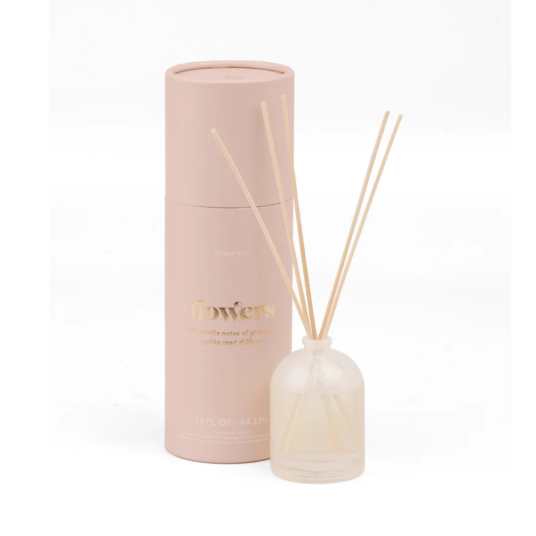 A reed diffuser set with a pale pink cylindrical bottle, a frosted glass bottle with natural reeds, and gold lettering, shown on a clean white background.