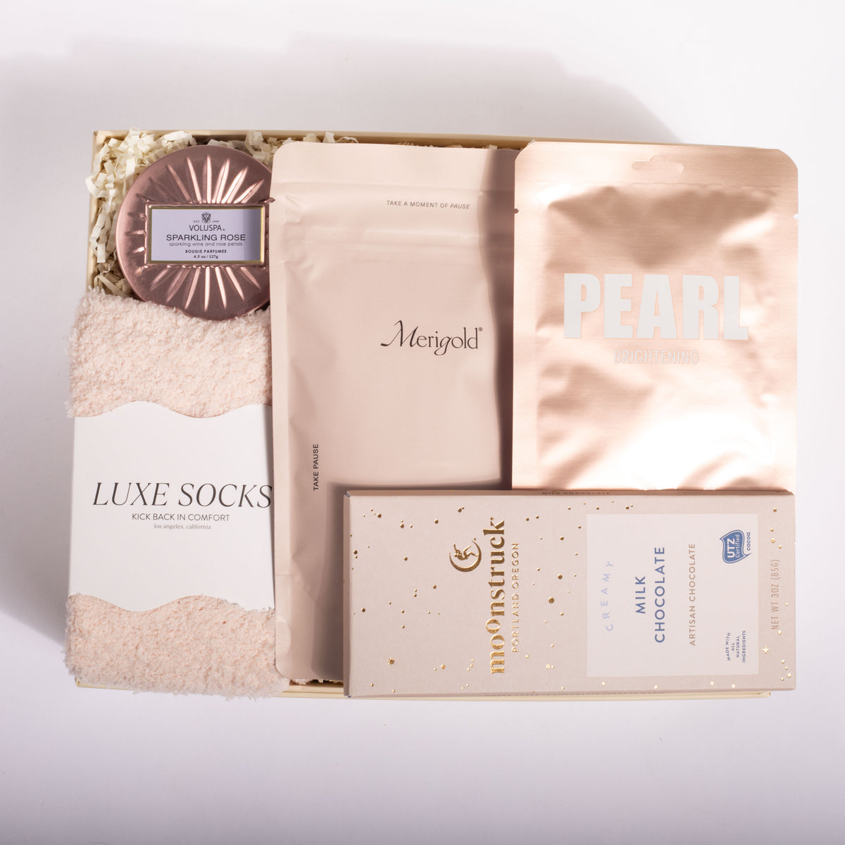 A curated gift box set containing a plush beige sweater, a round metallic rose gold soap, a package of Meriygold pouches, a package of Pearl Chocolate, and a box of Moonstruck Milk Chocolate, arranged in a box with shredded paper.